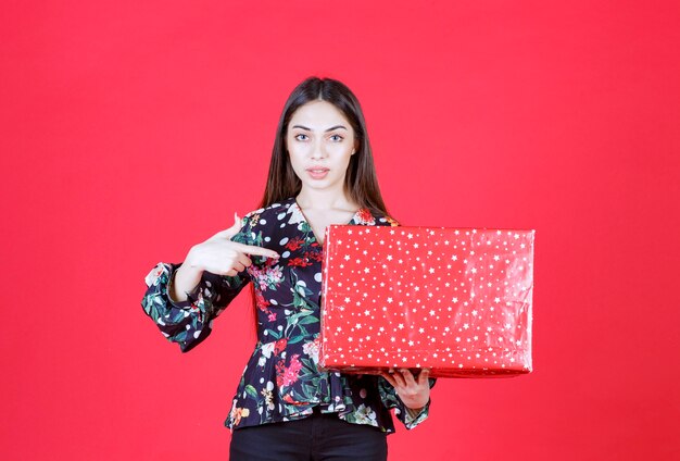 mujer con camisa floral sosteniendo una caja de regalo roja con puntos blancos.