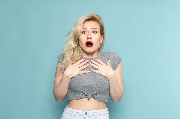 Mujer en camisa gris y jeans azul brillante posando con expresión asustada