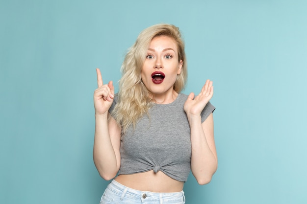 Mujer en camisa gris y jeans azul brillante posando con expresión de sorpresa