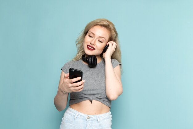 Mujer en camisa gris y jeans azul brillante sosteniendo un teléfono escuchando música
