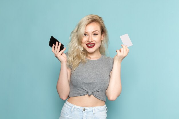 Mujer en camisa gris y jeans azul brillante sosteniendo teléfono y tarjeta blanca