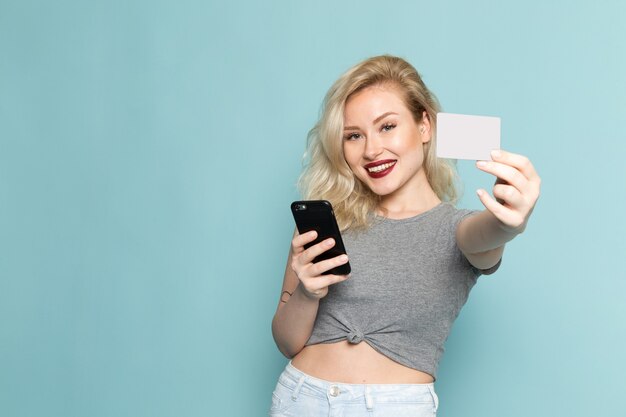 Mujer en camisa gris y jeans azul brillante sosteniendo teléfono y tarjeta blanca