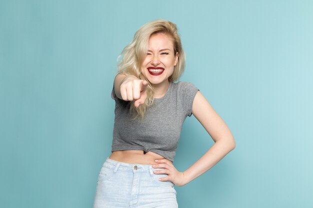 mujer en camisa gris y pantalones vaqueros azules brillantes posando con una sonrisa y expresión encantada