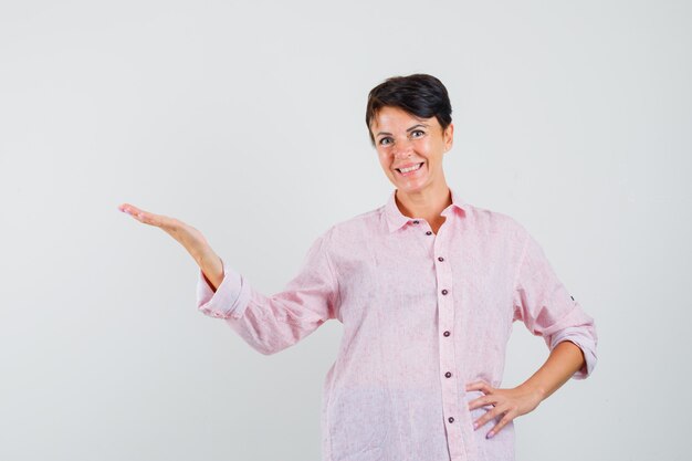 Mujer en camisa rosa dando la bienvenida o mostrando algo y mirando alegre, vista frontal.