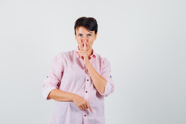 Mujer con camisa rosa mostrando gesto de silencio y mirando con cuidado, vista frontal.