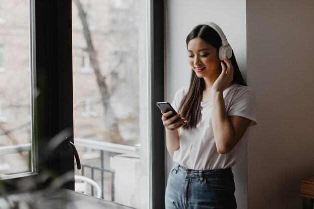 Mujer en camiseta blanca y jeans posa junto a la ventana con el teléfono en las manos Chica escuchando música en los auriculares