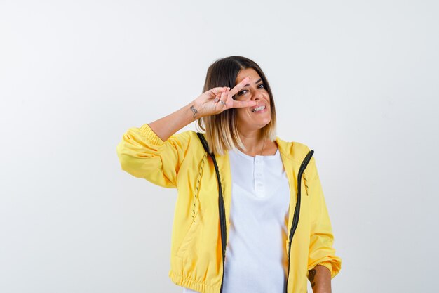 Mujer en camiseta, chaqueta que muestra el signo V cerca del ojo y luciendo alegre, vista frontal.