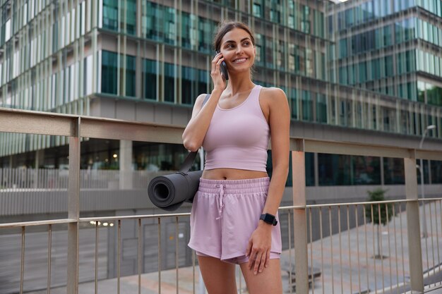 mujer en camiseta sin mangas y pantalones cortos hace una llamada telefónica mientras camina hacia el estadio al aire libre para practicar yoga para relajarse después de un día estresante consulta sobre ejercicios de sesión de entrenamiento llamando a alguien