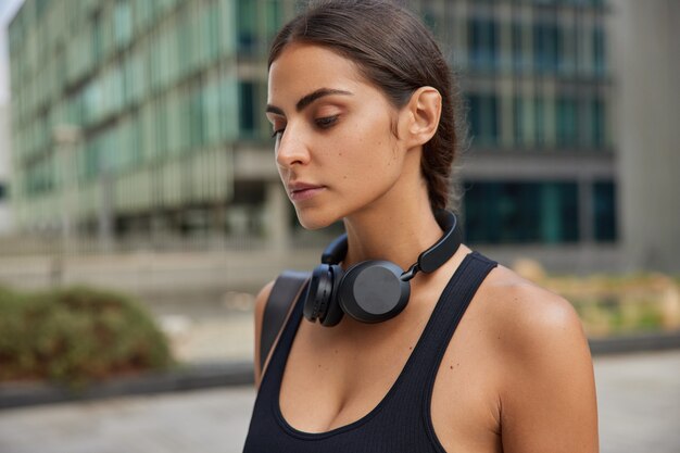 Mujer en camiseta negra usa auriculares inalámbricos mientras entrena al aire libre enfocado hacia abajo piensa en algo en el centro de la ciudad contra borrosa