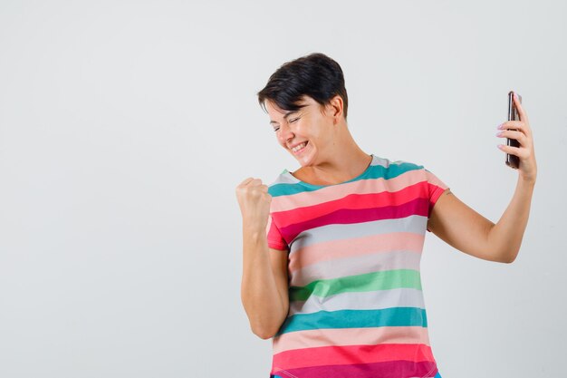 Mujer en camiseta a rayas que muestra el gesto del ganador, sosteniendo el teléfono móvil, vista frontal.