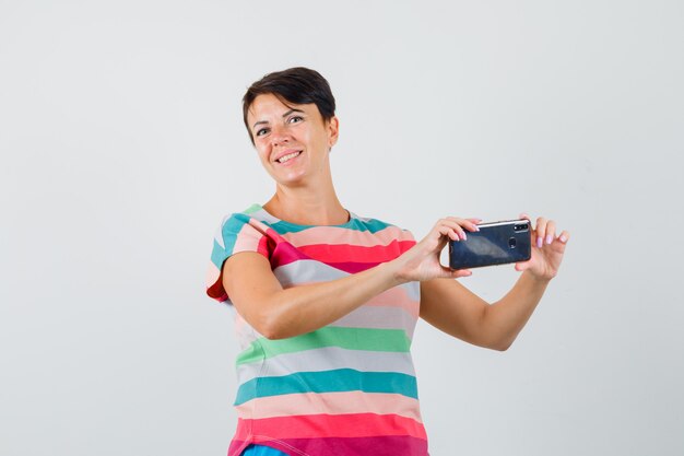 Mujer en camiseta a rayas tomando fotos en el teléfono móvil y mirando alegre, vista frontal.