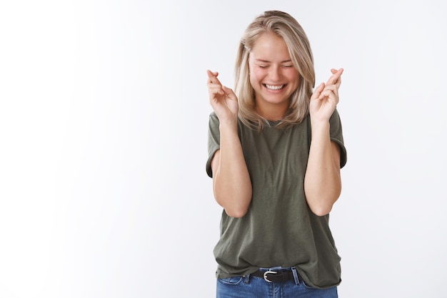 Mujer en camiseta verde oliva ojos cerrados sonriendo emocionado y divertido sosteniendo los dedos cruzados para la buena suerte como desear aganst blanco