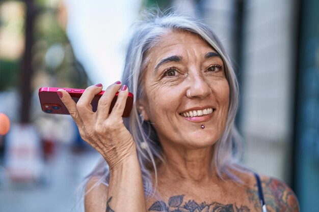 Mujer canosa de mediana edad sonriendo confiada escuchando un mensaje de audio por el teléfono inteligente en la calle