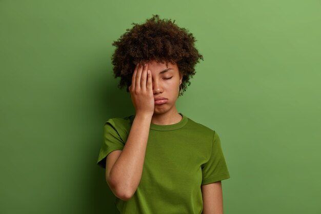 Mujer cansada agotada que hace cara con la palma de la mano, tiene problemas de salud, expresión de disgusto con sueño, suspiros de cansancio, usa camiseta verde informal, posa en interiores. Sentirse fatigado y con exceso de trabajo.