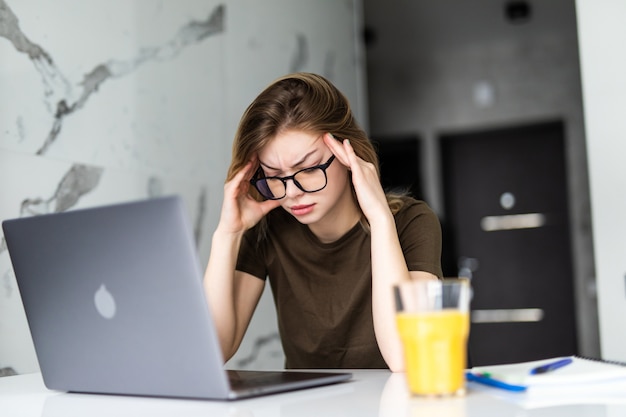 Mujer cansada agotada que trabaja usando en la computadora portátil sosteniendo la cabeza en la mano sentado en la cocina en casa