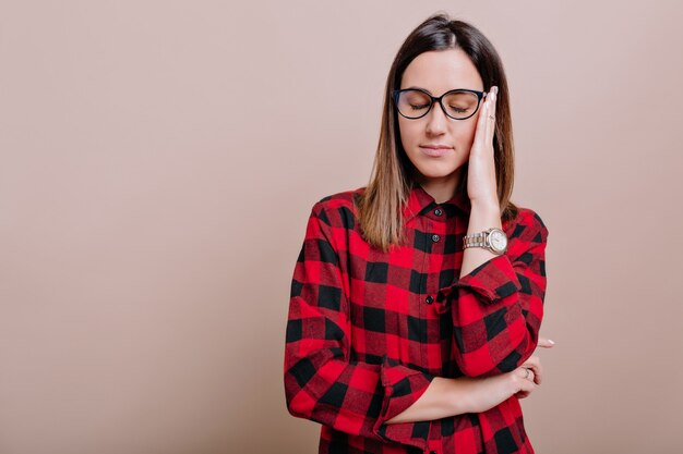 Mujer cansada con cabello oscuro y ojos cerrados lleva gafas sostiene la mano cerca de la cara en la pared aislada. La mujer con dolor de cabeza posa en la pared beige