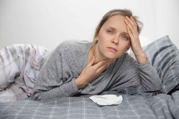 Mujer cansada enferma en la cama, con dolor de garganta, mirando a la cámara