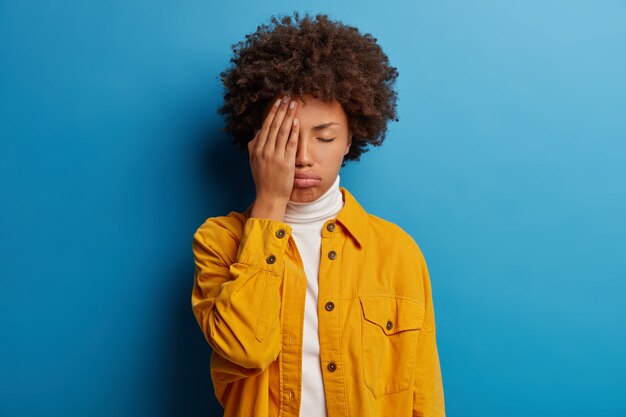 Mujer cansada y harta cubre la mitad de la cara, mantiene los ojos cerrados, suspira de cansancio, se siente agotada para trabajar sin descansar, posa en el estudio contra la pared azul