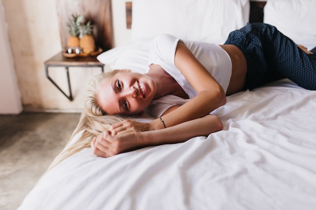 Mujer cansada en pantalones oscuros en sábana con sonrisa. Filmación en interiores de una linda dama rubia posando en la cama.