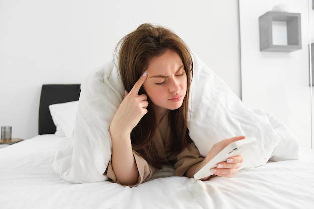 Foto gratuita mujer con cara confusa despertándose por la mañana y revisando su mensaje de lectura de teléfono móvil con