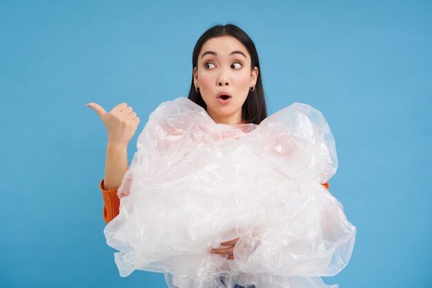 Foto gratuita mujer con cara sorprendida sosteniendo residuos de reciclaje de plástico y apuntando a la izquierda con emoción asombrada azul