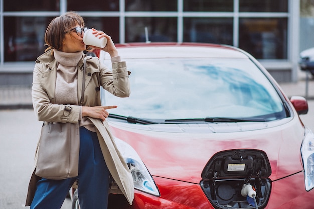 Mujer cargando un coche eléctrico en la gasolinera eléctrica y tomando café