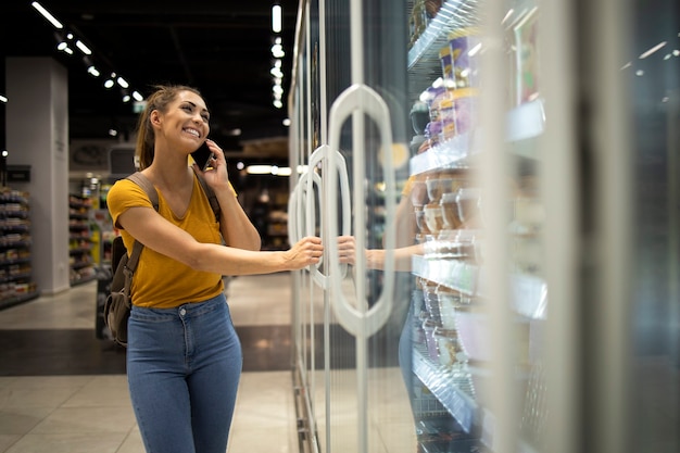 Mujer con carrito de compras abriendo nevera para llevar comida en la tienda mientras habla por teléfono