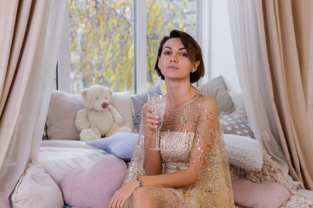 Mujer en casa acogedora habitación con vestido de noche brillante de Navidad, sosteniendo una copa de champán