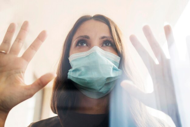 Mujer en casa en cuarentena con máscara médica detrás de la ventana