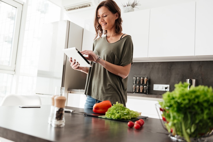 Foto gratuito mujer casual contenta usando tableta mientras está de pie cerca de la mesa en la cocina