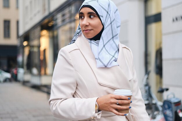 Mujer casual joven árabe en hijab tomando café durante el paseo por la calle de la ciudad