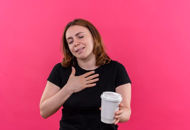 Mujer casual joven complacida sosteniendo una taza de café de plástico con la mano en el pecho con los ojos cerrados en el espacio rosa aislado con espacio de copia