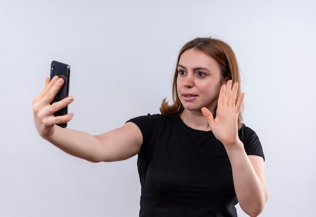 Mujer casual joven complacida sosteniendo el teléfono móvil y gesticulando hola al teléfono en un espacio en blanco aislado