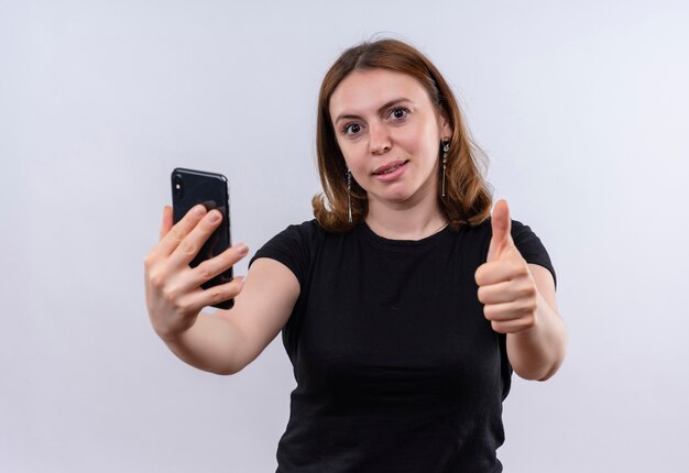 Mujer casual joven confiada que estira el teléfono móvil y que muestra el pulgar hacia arriba en el espacio en blanco aislado
