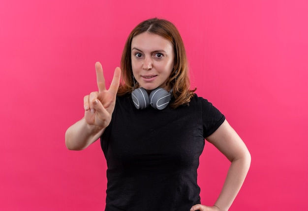 Mujer casual joven confiada que lleva puestos los auriculares en el cuello haciendo el signo de la paz en el espacio rosado aislado