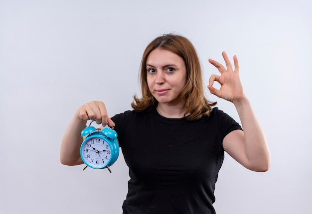 Mujer casual joven confiada que sostiene el reloj de alarma y que hace la muestra aceptable en el espacio en blanco aislado