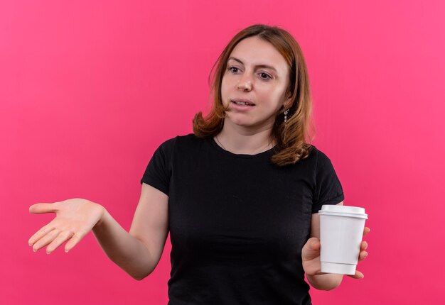 Mujer casual joven confundida que sostiene la taza de café plástica y que muestra la mano vacía en el espacio rosado aislado