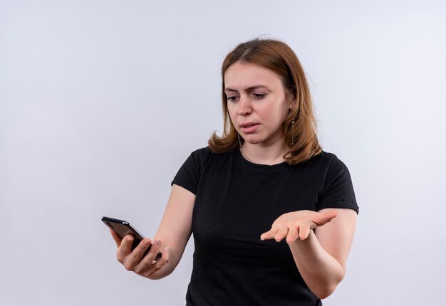 Mujer casual joven confundida que sostiene el teléfono móvil y que muestra la mano vacía en el espacio blanco aislado con el espacio de la copia