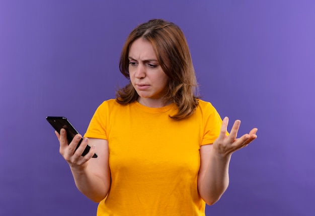 Foto gratuita mujer casual joven confundida que sostiene el teléfono móvil y que muestra la mano vacía y que la mira en el espacio púrpura aislado