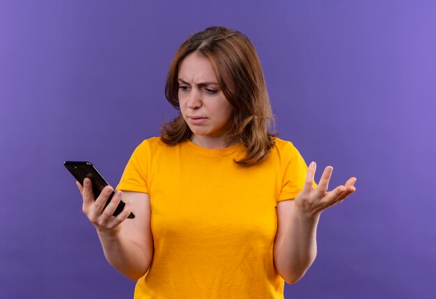 Mujer casual joven confundida que sostiene el teléfono móvil y que muestra la mano vacía y que la mira en el espacio púrpura aislado