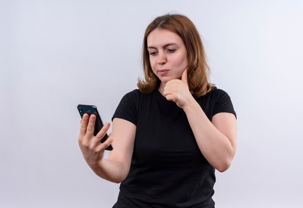 Mujer casual joven pensativa que sostiene el teléfono móvil y que pone la mano debajo de la barbilla en la pared blanca aislada con el espacio de la copia