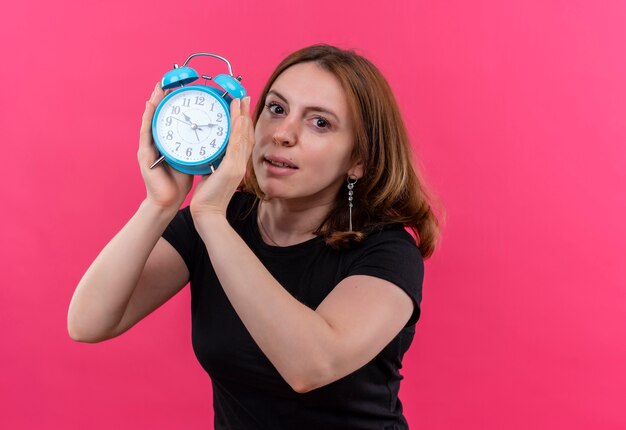 Mujer casual joven que sostiene el despertador y en la pared rosada aislada con el espacio de la copia