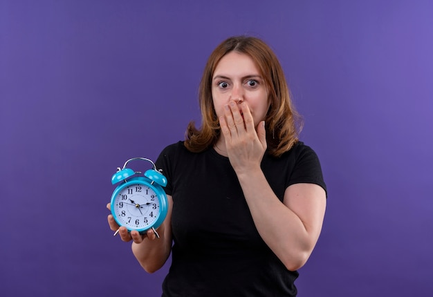 Mujer casual joven sorprendida que sostiene el reloj de alarma con la mano en la boca en la pared púrpura aislada con el espacio de la copia