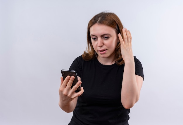 Mujer casual joven sorprendida sosteniendo el teléfono móvil y mirándolo con la mano cerca de la cabeza en la pared blanca aislada con espacio de copia