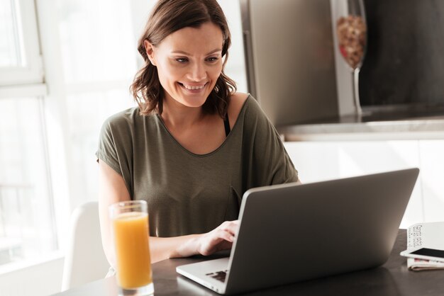 Mujer casual sonriente que usa la tableta junto a la mesa en la cocina