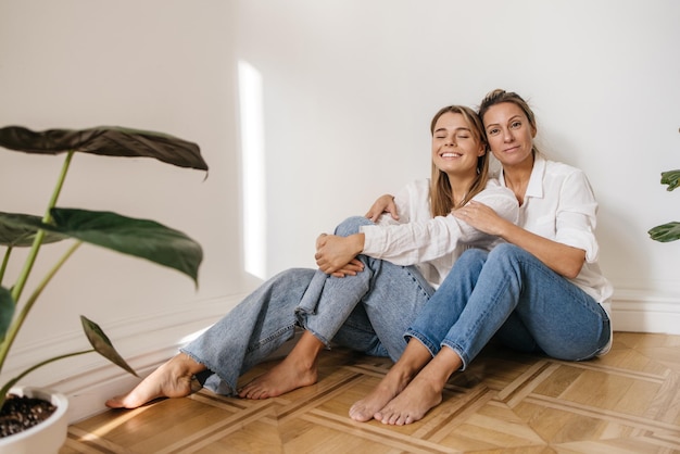 Foto gratuita una mujer caucásica adulta abraza a una joven sentada a su lado en el suelo contra una pared blanca. estilo de vida, concepto de belleza femenina