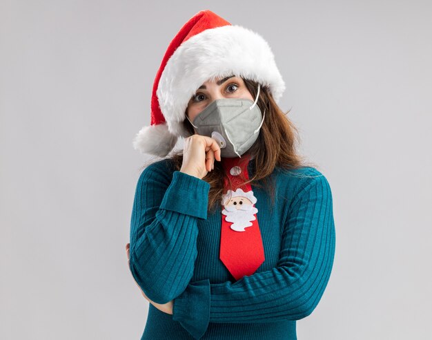 Mujer caucásica adulta ansiosa con gorro de Papá Noel y corbata de santa con máscara médica aislada en la pared blanca con espacio de copia