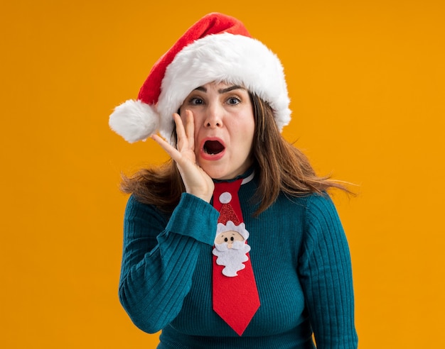 Mujer caucásica adulta ansiosa con gorro de Papá Noel y corbata de santa sostiene la mano cerca de la boca llamando a alguien aislado en la pared naranja con espacio de copia