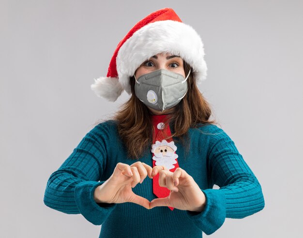 Mujer caucásica adulta complacida con gorro de Papá Noel y corbata de santa con máscara médica gesticulando signo de corazón aislado en la pared blanca con espacio de copia