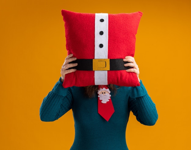 Foto gratuita mujer caucásica adulta con corbata de santa sosteniendo una almohada decorada delante de la cara aislada sobre fondo naranja con espacio de copia